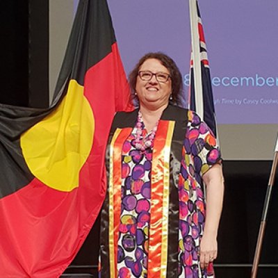 Lorelle Holland wearing a colourful dress and a black, red and yellow sash. She is standing in front of an Aboriginal, Australian, and Torres Strait Islands flag.